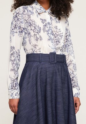 Femme portant un chemisier blanc à manches longues avec un imprimé floral bleu et une jupe bleu marine taille haute avec une large ceinture et une boucle.