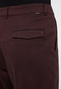 Burgundy trousers with a textured fabric, slim fit, and a back pocket featuring a split design. Branded tag is visible on the waistband.