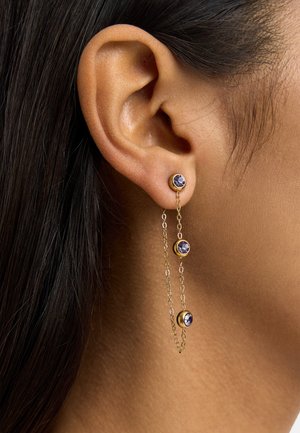 Złote kolczyki w formie łańcuszka z trzema małymi fioletowymi kamieniami, noszone na uchu osoby o ciemnych włosach i średnim odcieniu skóry.