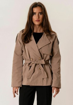 Hosszú barna hajú nő bézs kétsoros gombolású, öves kabátban és fekete nadrágban, semleges háttér előtt állva.