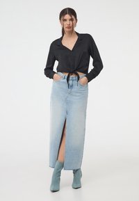 Camicetta nera corta con bottoni e un nodo in vita, abbinata a una gonna maxi in denim azzurro chiaro con uno spacco frontale e stivali alla caviglia color teal.
