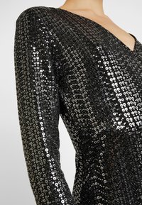 Svart paljettklänning med långa ärmar, med en djup V-ringning och en texturerad yta skapad av otaliga cirkulära paljetter.