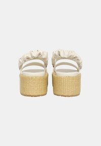 Sandali con tacco alto con una parte superiore circolare beige in tessuto, dotati di un design arricciato e una suola piatta intrecciata di colore marrone chiaro.