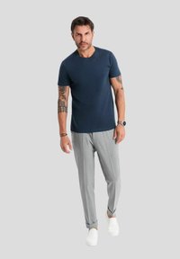 T-shirt navy a girocollo, pantaloni slim fit grigi chiari e sneaker bianche. Tatuaggi visibili sull'avambraccio sinistro; texture del tessuto liscia.