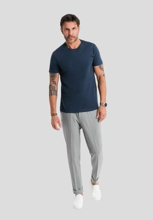 Navy crew-neck t-shirt, slim-fit light gray trousers, and white sneakers. Tattoos visible on left forearm; smooth fabric texture.