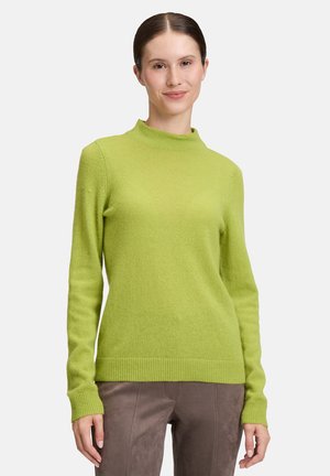 Femme portant un pull à manches longues vert citron et un pantalon marron, se tenant contre un fond clair, souriant légèrement.