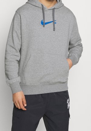 Man draagt een grijze hoodie met blauw Nike-logo en zwarte broek met wit Nike-logo, poseert met één hand in de zak van de hoodie.