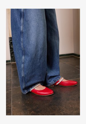 Sapatilhas de couro vermelho com ponta arredondada e uma tira de destaque prateada. Usadas com jeans azuis de perna larga, apresentando uma bainha desfiada.