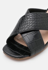 Sandale noire avec un design texturé imitant le crocodile, dotée d'une large sangle entrecroisée et d'une pointe ouverte, reposant sur une semelle plate beige.