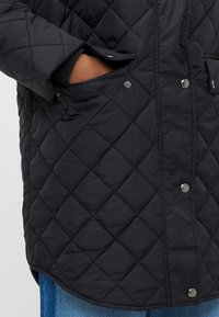 Črna prešita jakna z diamantnim vzorcem, z zapenjanjem na pritisne gumbe in stranskimi žepi. Mehka tkanina in rahlo ukrivljen rob na spodnji strani.