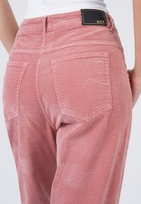 Roze ribfluwelen broek met een geribbelde textuur, voorzien van twee achterzakken en een zwart etiket met "M.O.D" op de tailleband.