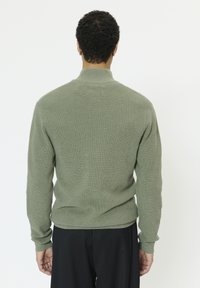 Pull vert en maille à manches longues et col montant, présentant un motif texturé. Porté avec un pantalon noir, vu de dos.