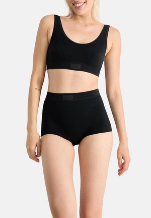 Woman wearing black high-waist briefs and black crop top, standing against plain light background with a slight smile.