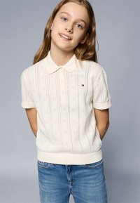 Polo branco de malha, com mangas curtas e colarinho canelado, apresentando um padrão texturizado e um pequeno logotipo no peito. Combinado com jeans azuis.
