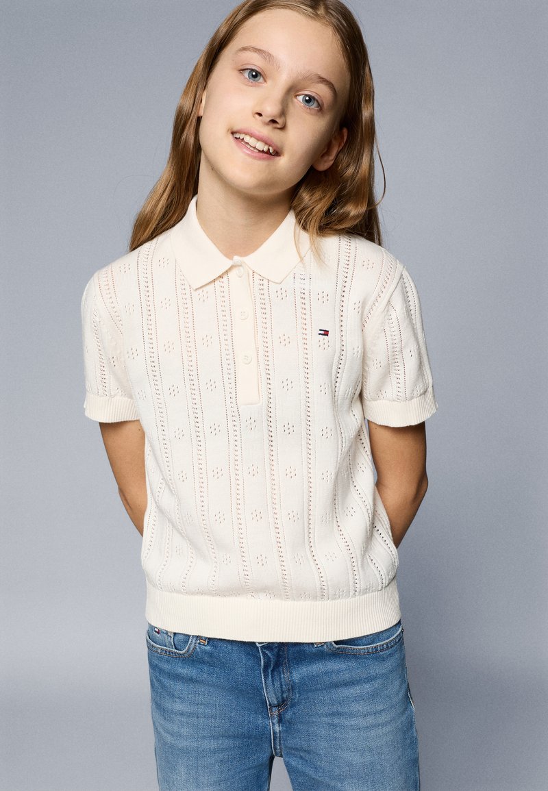 Polo branco de malha, com mangas curtas e colarinho canelado, apresentando um padrão texturizado e um pequeno logotipo no peito. Combinado com jeans azuis.