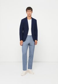 Granatowa marynarka, biała koszulka i jasnoniebieskie spodnie o slim fit. Strukturalna tkanina, minimalistyczny design i jasne buty wsuwane.