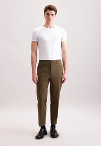 Pantaloni verde oliva a taglio aderente realizzati in tessuto liscio, indossati con una t-shirt bianca a girocollo. Presentano tasche laterali e pieghe frontali.