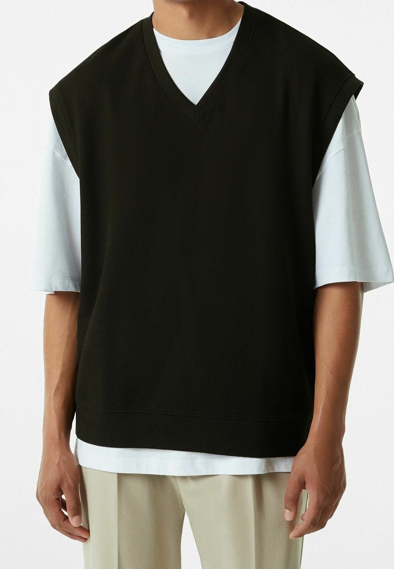 Černá vesta s krátkými rukávy a výstřihem do V z texturované látky, nošená přes bílou košili, kombinovaná se světlými kalhotami.