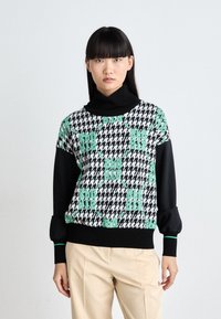 Mustavalkoinen houndstooth-kuvioinen neule, jossa on vihreät tekstiakcentit. Varustettu korkealla kauluksella ja puffihihoilla, ja sen alaosa on kontrastina musta.