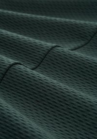 Sötétzöld textúrált szövet, gyémántmintázattal, finom ráncokkal és varratokkal, amelyek kiemelik lélegző és könnyű anyagát.