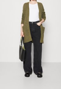 Olivgrön stickad cardigan med öppen front, två fickor, bärs över en vit skjorta; tillsammans med svarta vida jeans och svarta slip-on skor.