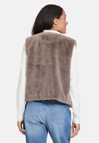 Frau trägt eine ärmellose taupefarbene Kunstpelzweste über einem weißen Langarmoberteil und hellblauen Jeans, mit dem Rücken zur Kamera gewandt.