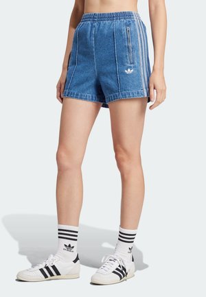 Henkilö pukeutunut sinisiin farkku-Adidas-shortseihin, joissa on kolme valkoista raitaa ja logo, valkoiset Adidas-sukat sekä valkoiset lenkkarit mustilla raidoilla.