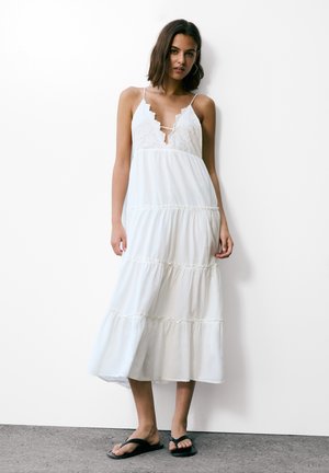 Robe maxi blanche avec des détails en dentelle, des bretelles spaghetti, un design en plusieurs niveaux et un ourlet volé. Associée à des tongs noires sur un sol gris.