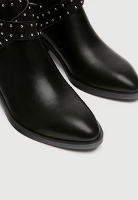 Botas de tobillo de cuero negro con puntera afilada, que cuentan con una correa decorativa adornada con pequeños tachuelas de plata. Superficie lisa y brillante.