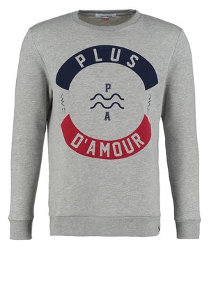 Mann trägt grauen Pullover mit dem Schriftzug „Plus d'amour“, hell ausgewaschene Jeans, weiße Slip-On-Schuhe und trägt einen braunen Rucksack über einer Schulter.