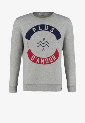 Mann trägt grauen Pullover mit dem Schriftzug „Plus d'amour“, hell ausgewaschene Jeans, weiße Slip-On-Schuhe und trägt einen braunen Rucksack über einer Schulter.