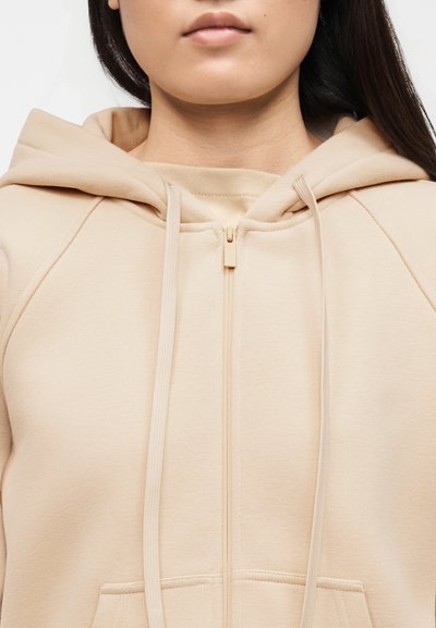 Κοντινό πλάνο ενός ατόμου που φοράει μια μπεζ ζακέτα με φερμουάρ, κουκούλα και κορδόνια, παρουσιάζοντας το στήθος και το κάτω μέρος του προσώπου.