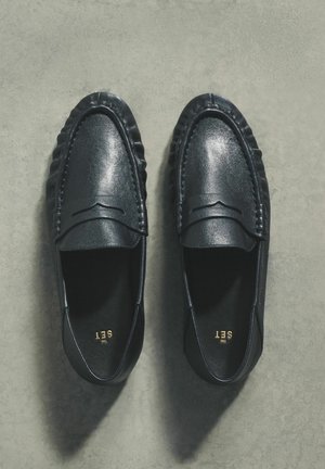 Melni ādas loaferi ar gludu tekstūru, tiem ir šūta detaļa gar augšdaļu un zems profils. Iekšpusē redzams zelta logotips.