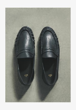 Svarta läderskor med slät struktur, med sydd detalj längs överdelen och låg profil. Insidan visar en logotyp i guld.