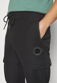Homme portant un pantalon décontracté noir avec taille à cordon, poche latérale zippée, poche cargo et sa main reposant dans la poche zippée.