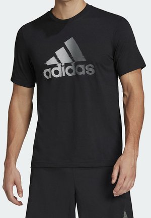 Mężczyzna w czarnej koszulce Adidas z srebrnym logo i czarnych szortach, stojący na jednolitym tle.
