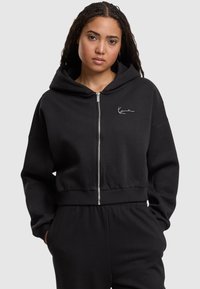 Felpa corta nera realizzata in misto cotone, con zip intera, grande cappuccio e un piccolo logo bianco sul lato sinistro del petto.