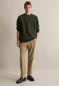 Dunkelgrüner strukturierter Pullover, beiger Cordhosen, schwarze Schuhe. Das Modell steht mit den Händen in den Taschen vor einem neutralen Hintergrund.