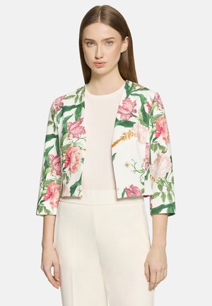 Femme portant une veste blanche courte avec un imprimé floral rose et vert sur un haut crème et un pantalon crème taille haute.