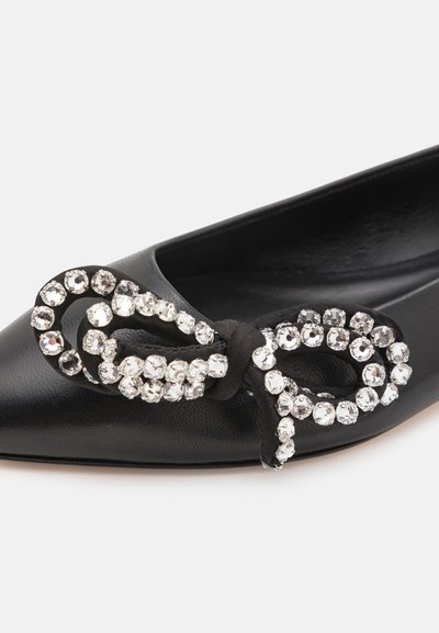 Zapato plano de cuero negro con punta afilada y un lazo decorativo negro adornado con strass transparentes dispuestos en un patrón circular.
