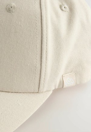 Cappello di stoffa beige con una forma strutturata, caratterizzato da un piccolo etichetta con logo e dettagli di cucitura visibili. Il materiale appare texture e resistente.