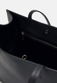 Intérieur d'un sac fourre-tout en cuir noir avec une poche zippée et un petit fermoir doré à l'intérieur.