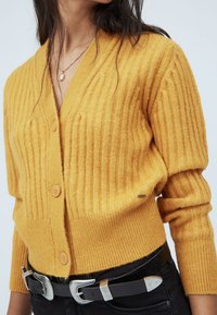 Kvinna i senapsgul ribbad cardigan med knappar, svart bälte med silverspänne och mörka byxor, som visar överkropp och nacke.