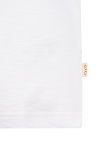 Gros plan sur un tissu blanc montrant la couture de l’ourlet et une petite étiquette beige avec un texte orange attachée sur le côté droit.