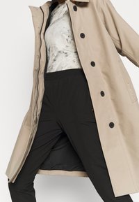 Abrigo largo beige con botones negros, llevado sobre una blusa con estampado blanco y gris y pantalones negros con cintura elástica.