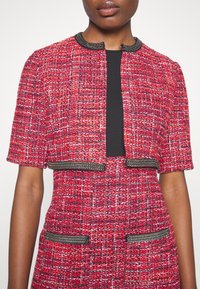 Femme portant une veste courte en tweed rouge et une robe avec un détail de chaîne noire au col et aux poches, par-dessus un top noir.