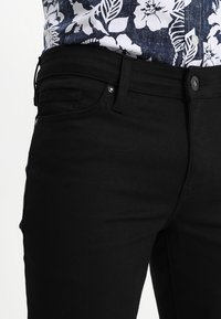 Svarta denimjeans med smal passform, med framfickor, knappstängning och stygnsdetaljer. Texturen verkar slät.