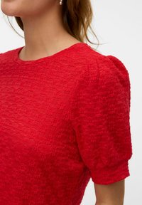 Maglietta rossa con texture, maniche corte a palloncino e scollatura rotonda, caratterizzata da un sottile motivo intrecciato su tutta la stoffa.