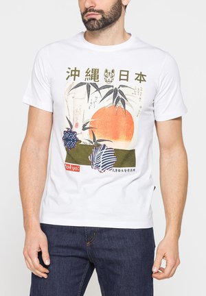 Uomo con maglietta bianca con caratteri giapponesi, sole arancione, foglie di bambù e fiori blu decorati, in piedi con jeans su sfondo semplice.
