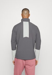 Grijze jas met hoge kraag, lichtgrijze accenten op de mouwen en een verticale lichtgrijze streep langs de achterkant. Gladde textuur, casual pasvorm.